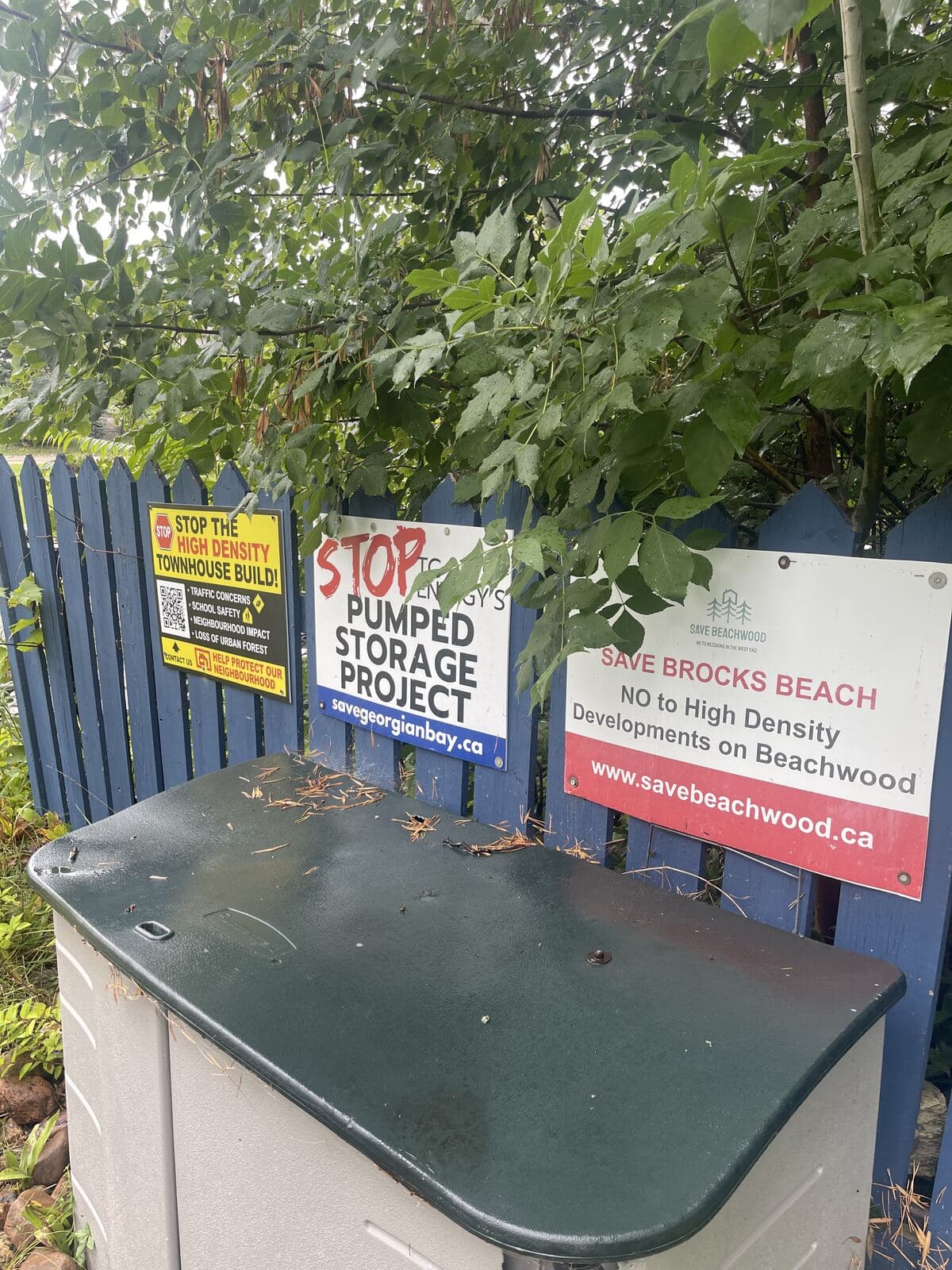Three residents' protest signs fixed to a blue picket fence in the west end of Wasaga Beach, reading Stop the high density townhouse build, Stop TC Energy's pumped storage project, and Save Brocks Beach.